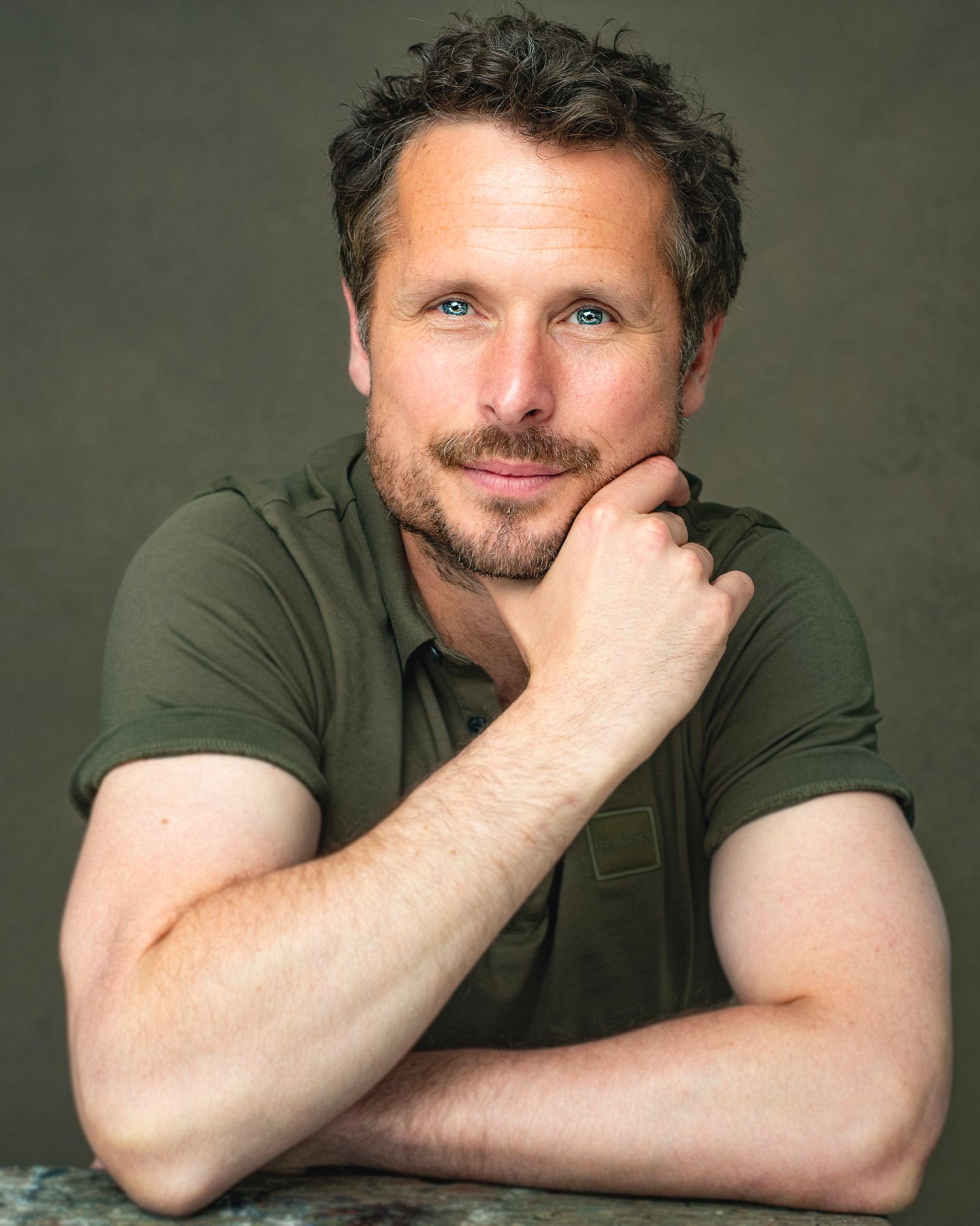 Professional portrait of someone in an olive green t-shirt with hand resting on chin.