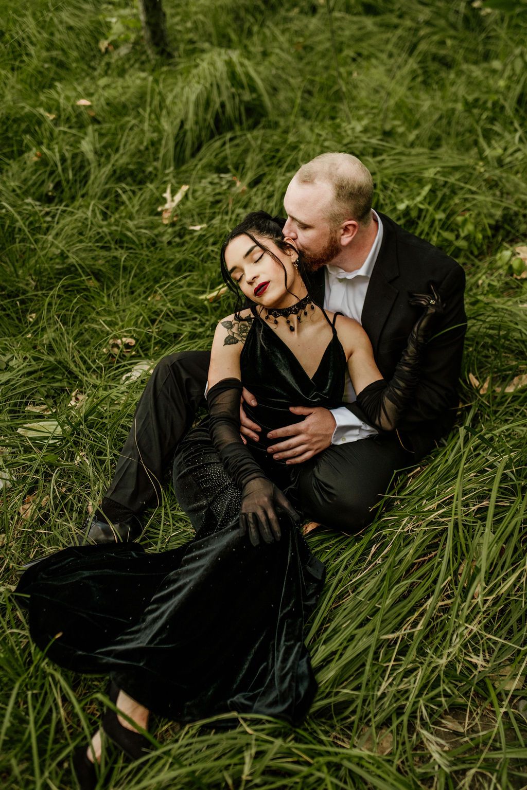 Romantic couple in formal attire embrace on grass in dramatic outdoor photo session.