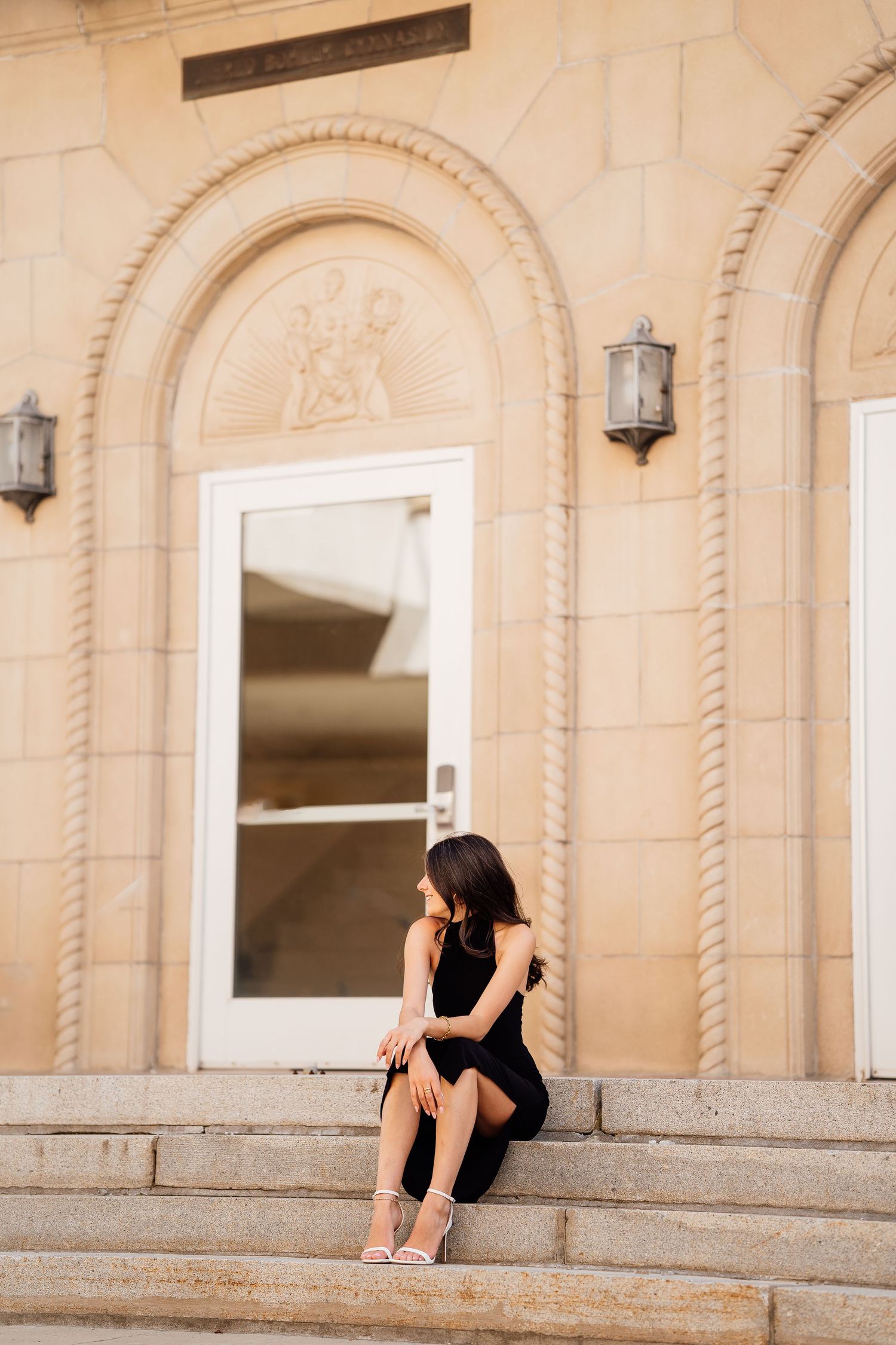 Person in black dress sitting elegantly on stone steps of building with ornate architecture.