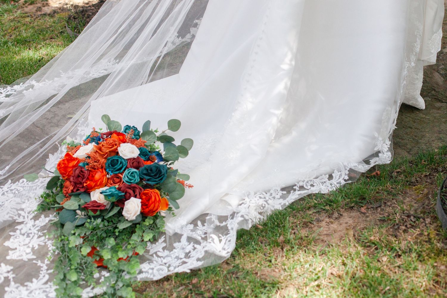 Detail of white wedding dress train with orange and teal floral accents on grass.