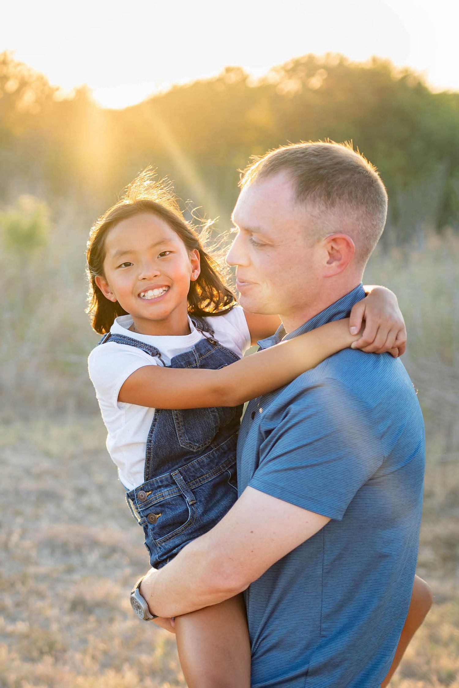 dad in blue shirt holding daughter in white shirt and denim overall with sun rays