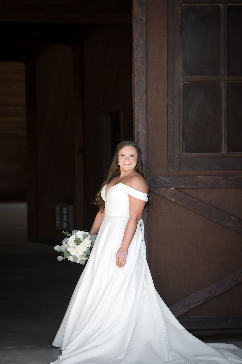 A bride in an elegant white gown poses in a rustic barn setting with dramatic lighting.