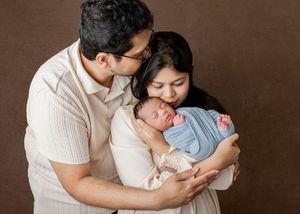 New parents tenderly hold their swaddled newborn baby in a warm studio portrait photo.