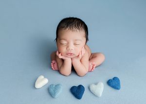 Peaceful newborn baby sleeping with chin on hands surrounded by blue and white felt hearts on a soft blue background.