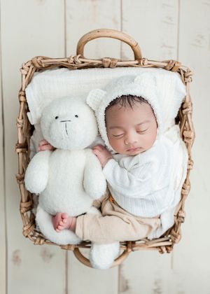 Sleeping newborn baby in bear bonnet nestled in wicker basket with white teddy bear, wrapped in soft white swaddle.