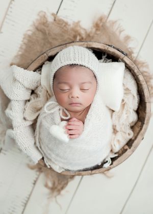 Sleeping newborn baby wrapped in cream knit swaddle and bonnet, nestled in a rustic wooden basket with soft fur lining.