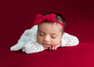 Sleeping newborn baby in white lace outfit with red bow headband on red background.