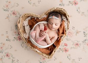 Newborn baby sleeping peacefully in a heart-shaped wicker basket with floral backdrop and rust-colored fabric.