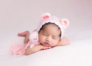 Sleeping newborn baby wearing a pink knit bear hat, cuddling a small pink stuffed animal on a soft white blanket.