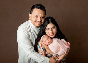A couple smiles warmly while cradling their sleeping newborn baby wrapped in pink in a professional studio portrait.