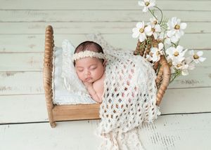Newborn baby sleeping peacefully in a tiny wooden bed with a white knit blanket and floral headband beside white flowers.