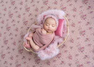 Newborn baby sleeping peacefully in a small basket with pink floral backdrop, wrapped in a soft pink swaddle and bonnet.