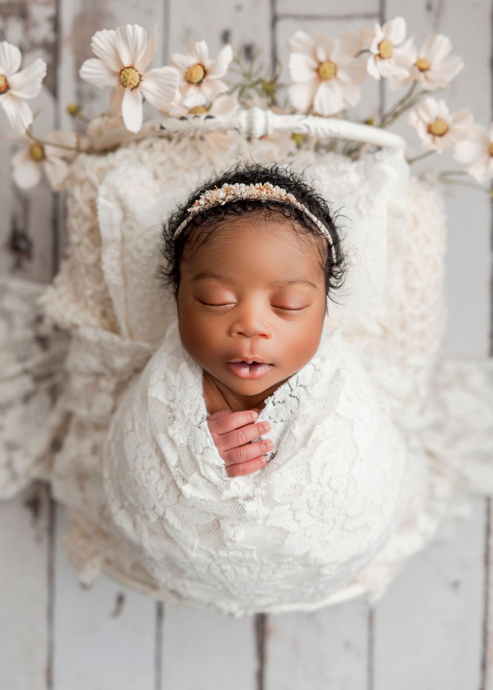 A sleeping newborn wrapped in white lace fabric surrounded by delicate white daisies in a dreamy photography setup.