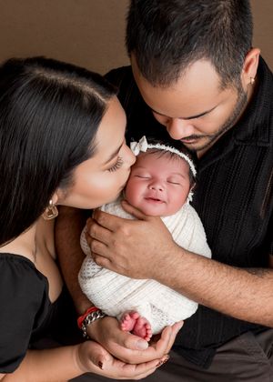 A newborn baby wrapped in white fabric is tenderly held and kissed by two adults wearing black against a brown backdrop.