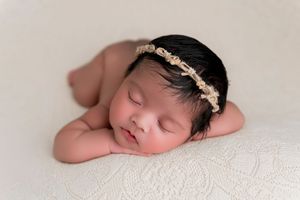 A sleeping newborn baby wearing a delicate gold leaf headband rests peacefully on white fabric.
