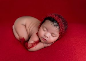 Sleeping newborn baby wearing red lace headband curled up on soft red backdrop during professional photo session.