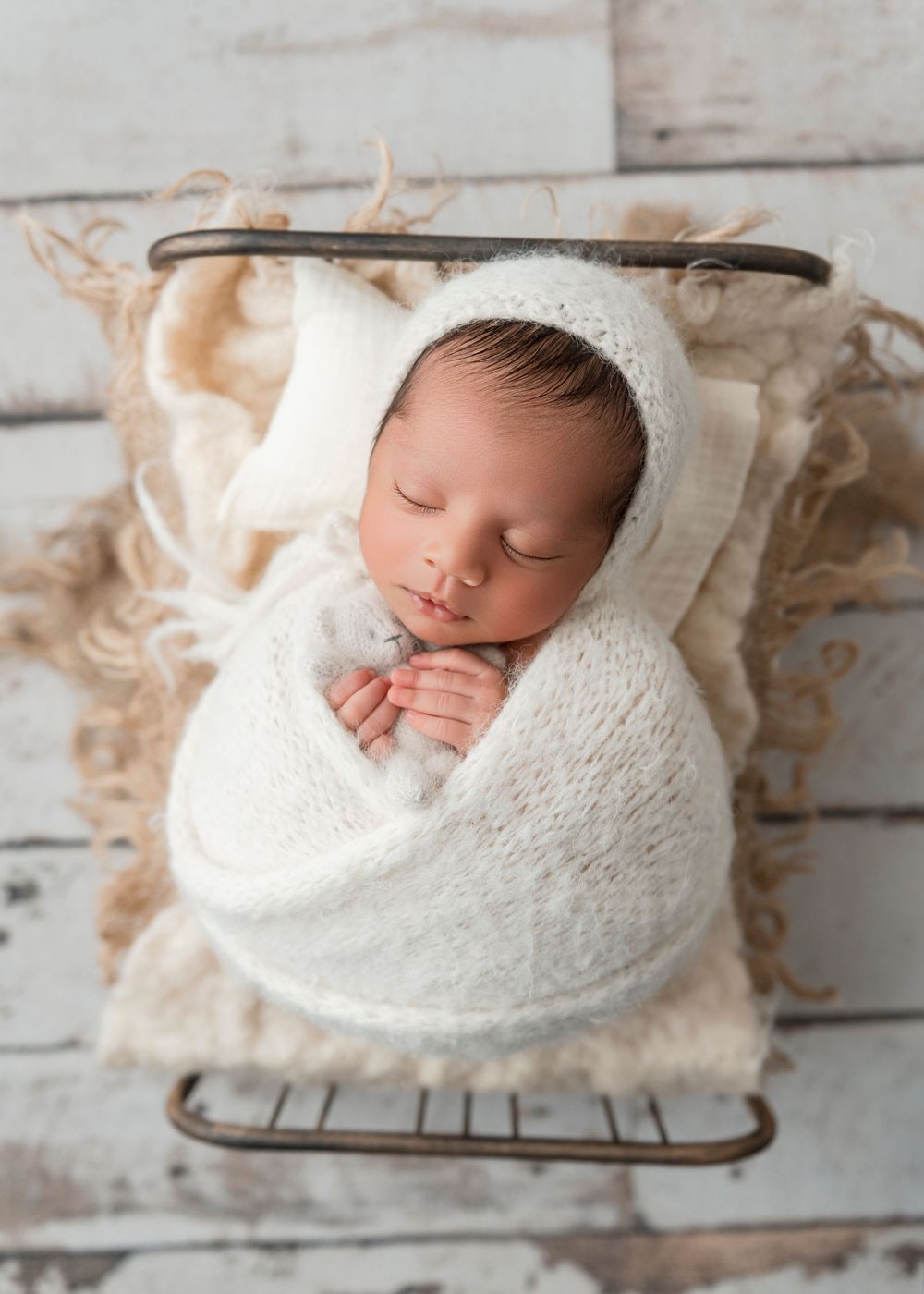 A sleeping newborn baby wrapped in a soft white knit blanket on a rustic wooden backdrop.