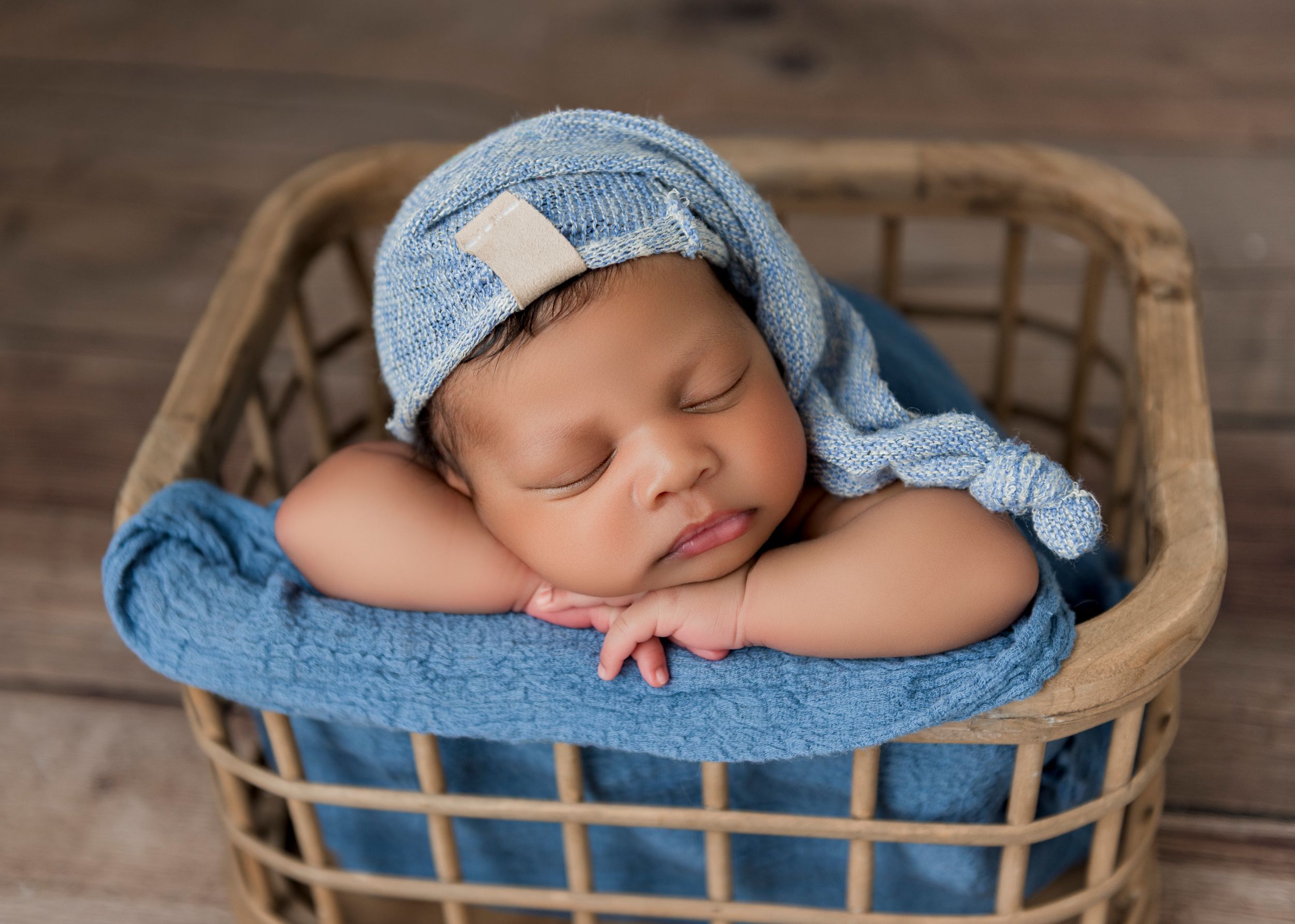 A sleeping newborn wearing a light blue hat rests peacefully in a woven basket on a wooden floor.