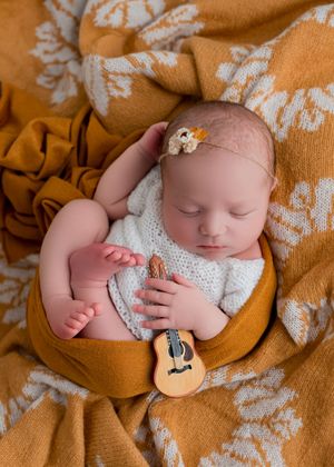 Sleeping newborn in white knit outfit holds tiny decorative guitar while resting on orange floral blanket.