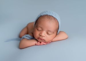 Peaceful sleeping newborn baby wearing light blue knit hat rests on soft blue fabric backdrop.
