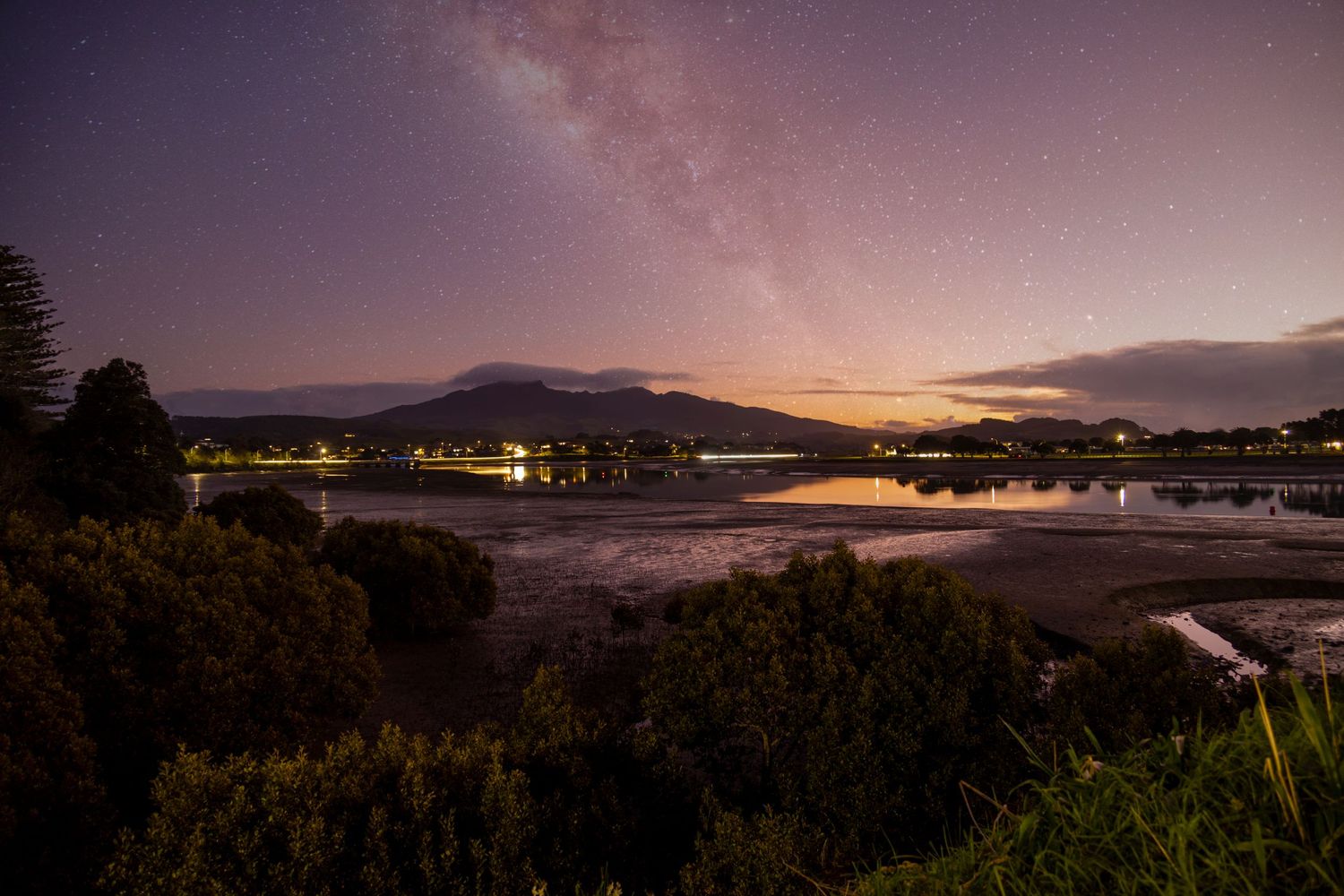 A star-streaked sky glows above Tauranga, with the Milky Way arching over calm waters and silhouetted hills.