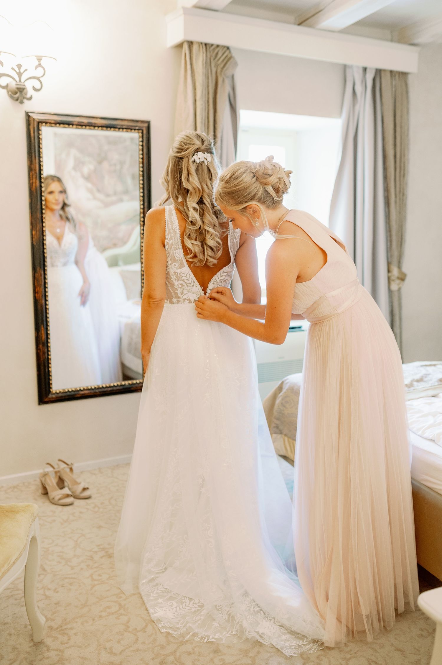 Two people in a bridal suite helping with the final adjustments of a white lace wedding gown.