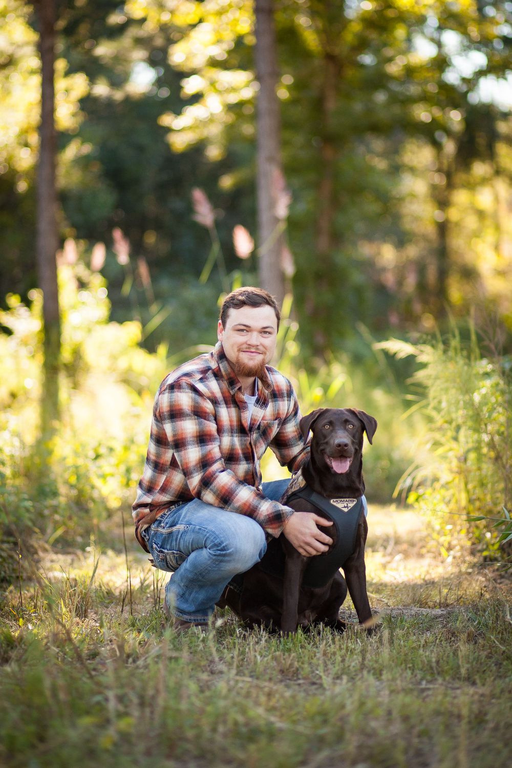 Person in plaid shirt kneeling beside black Labrador retriever on wooded path.