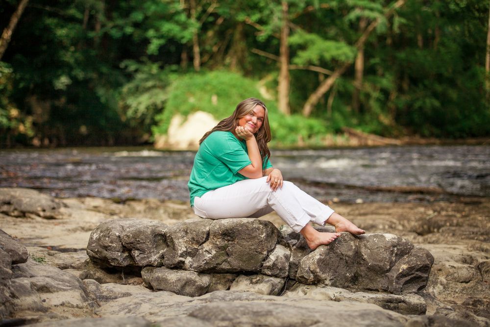 A contemplative figure sits on rocky outcrop by flowing water wearing green and white casual attire.