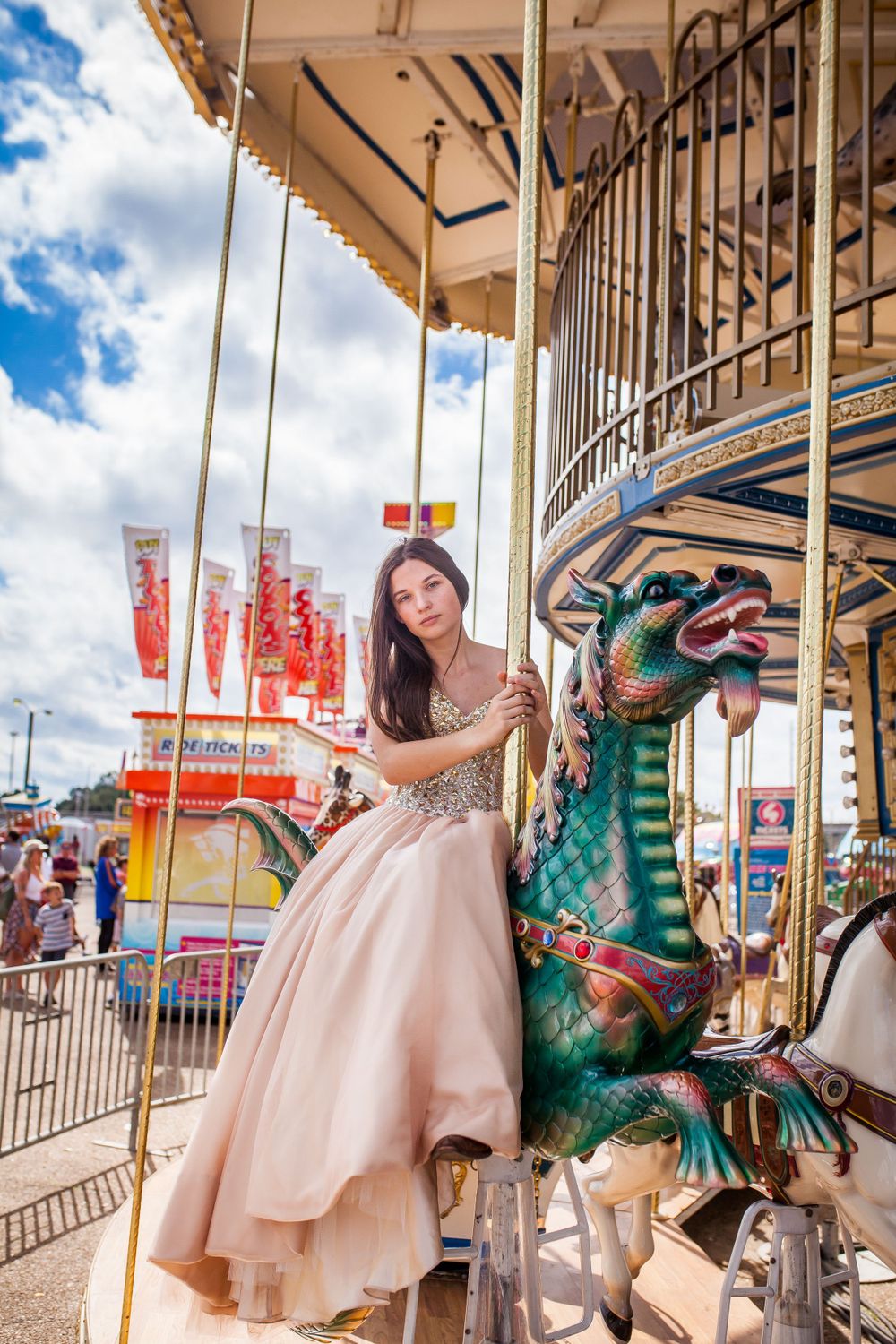 A person in a pink ballgown poses on a carousel horse at a carnival with blue skies and fair rides in background.