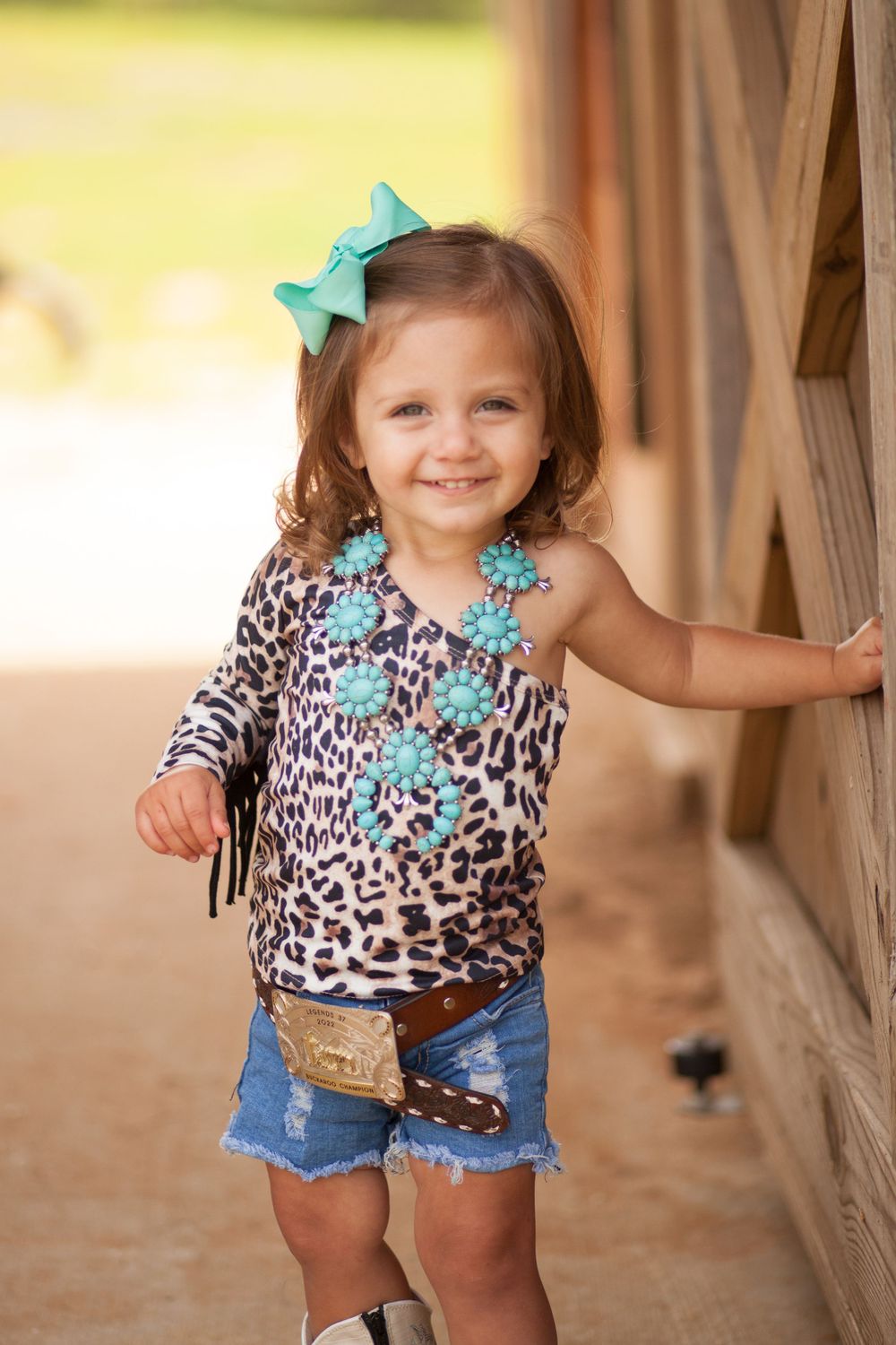 Child wearing leopard print top with turquoise bows and denim shorts stands by wooden wall.