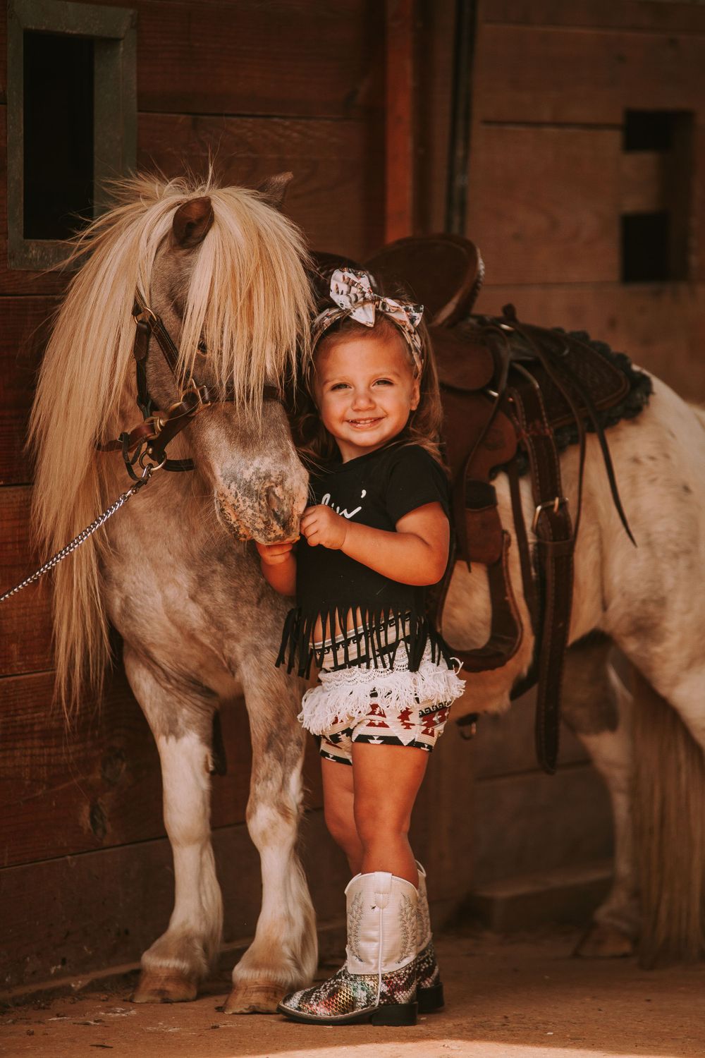 Child standing next to small pony in barn setting wearing western attire and boots.