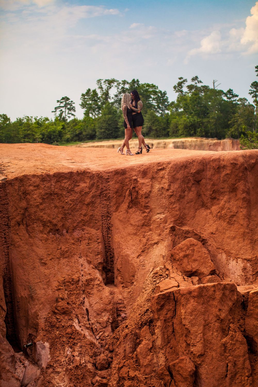 Two silhouettes stand at edge of dramatic red clay cliff during sunset.