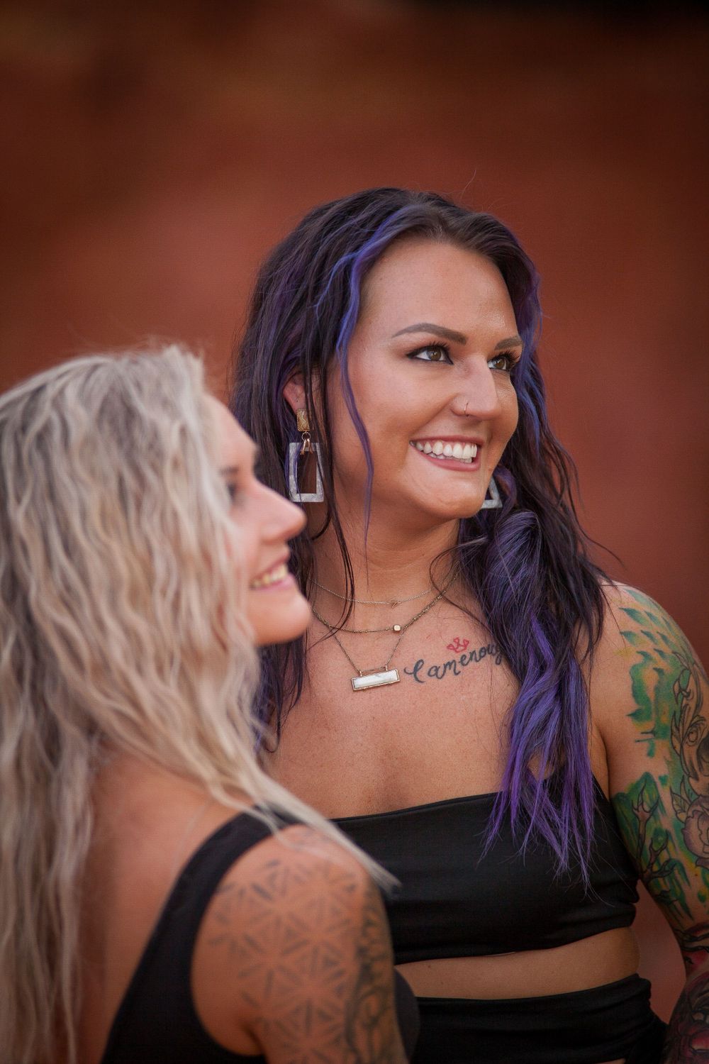 Two people with colorful hair and tattoos share joyful moment against rustic background.