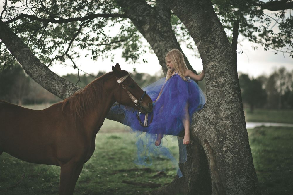 Woman in flowing blue dress stands near horse under large tree in dreamy scene.