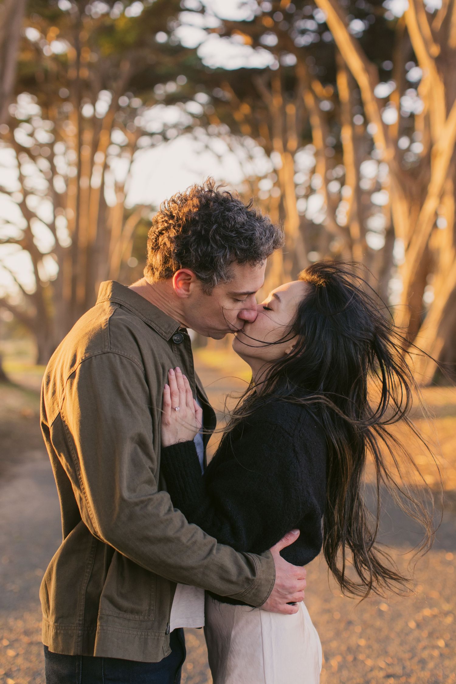 Intimate couple sharing a romantic kiss among tall cypress trees at sunset.