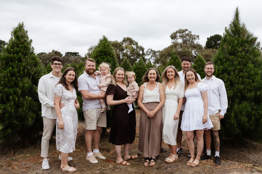 Extended family group photo taken outdoors at a Christmas tree farm in coordinated neutral outfits.