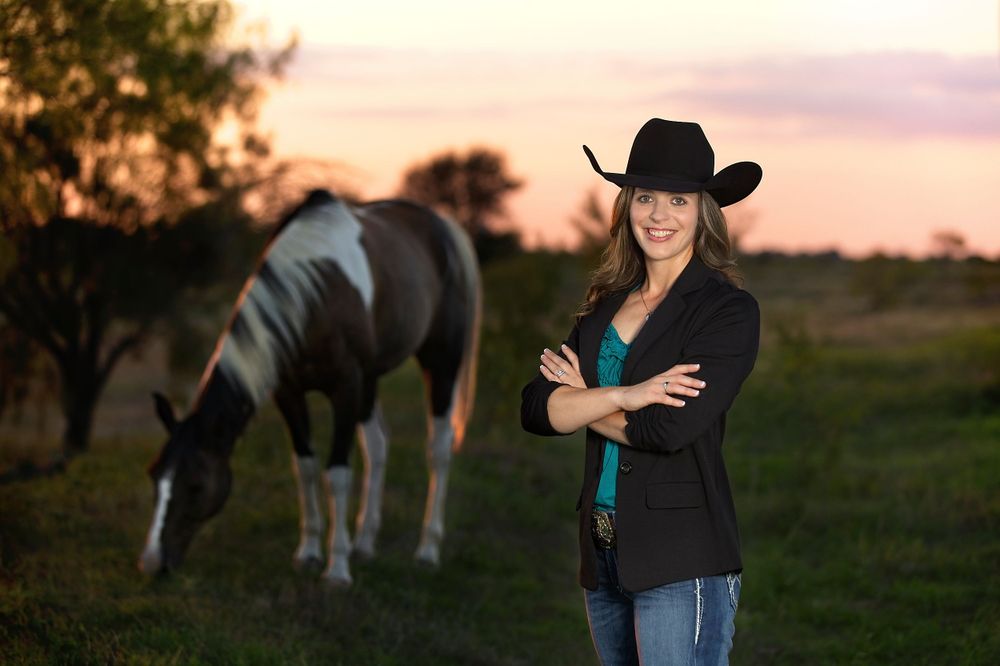 Woman in cowboy hat standing in field with grazing horse at sunset.