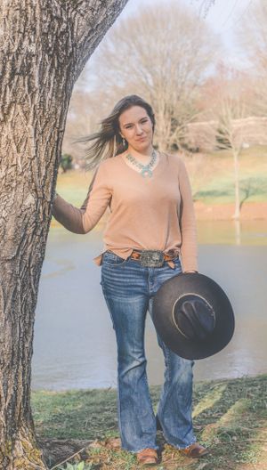 A woman wearing a peach colored top and jeans stands beside a tree trunk holding a black hat.