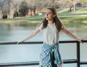 A person wearing a gingham shirt poses on a wooden railing by a lake.