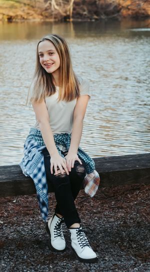 A person in casual attire sits on a wooden platform by a lake wearing white sneakers.