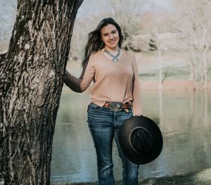 A woman in a peach sweater and jeans poses casually by a tree near a lake on a sunny day.