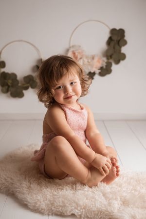 Toddler in pink dress sits on fluffy rug with decorative garland in background.