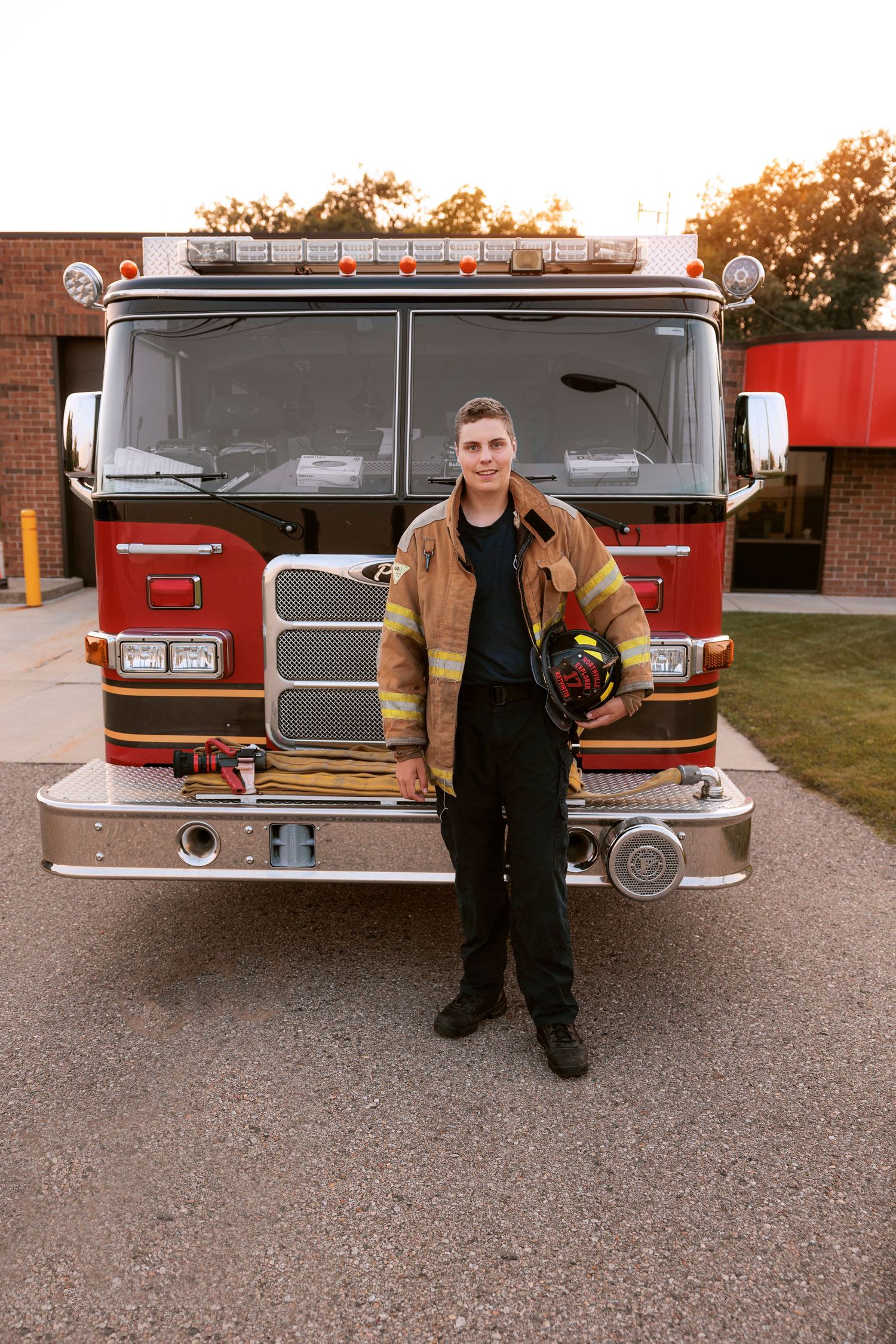 A firefighter stands proudly in front of a red fire engine wearing tan turnout gear and holding a helmet.