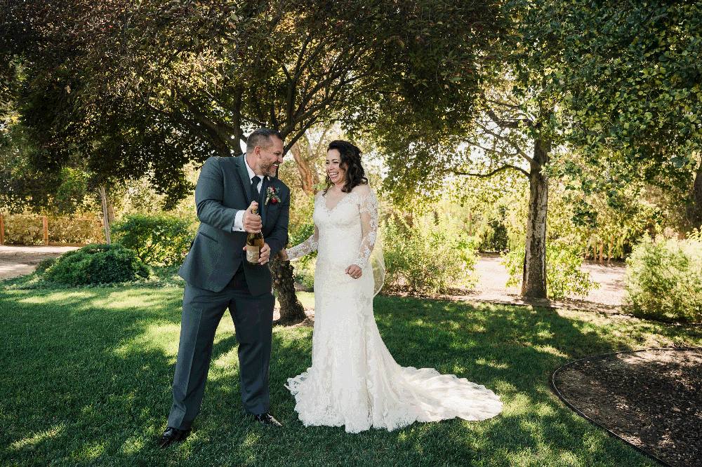 Newlyweds share champagne toast in sunlit garden setting.
