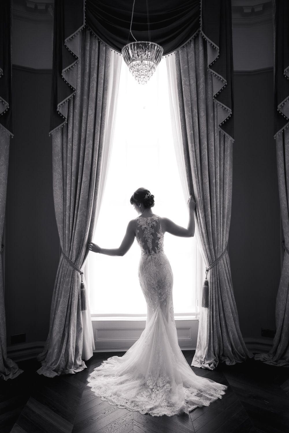 A silhouette of a bride in a lace gown standing by tall curtained windows with a crystal chandelier.