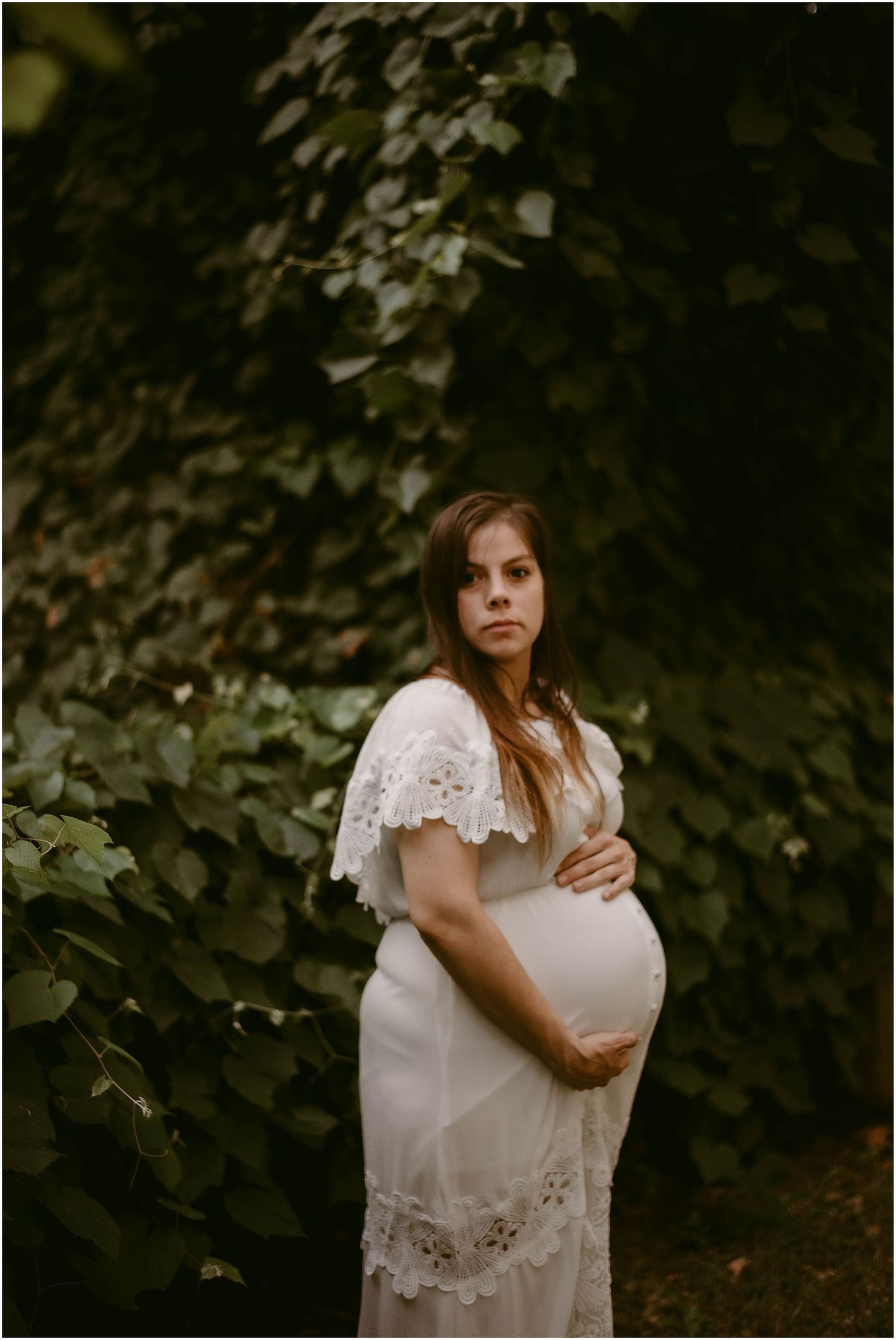 Moody maternity portrait of pregnant woman in white dress against dark green foliage.