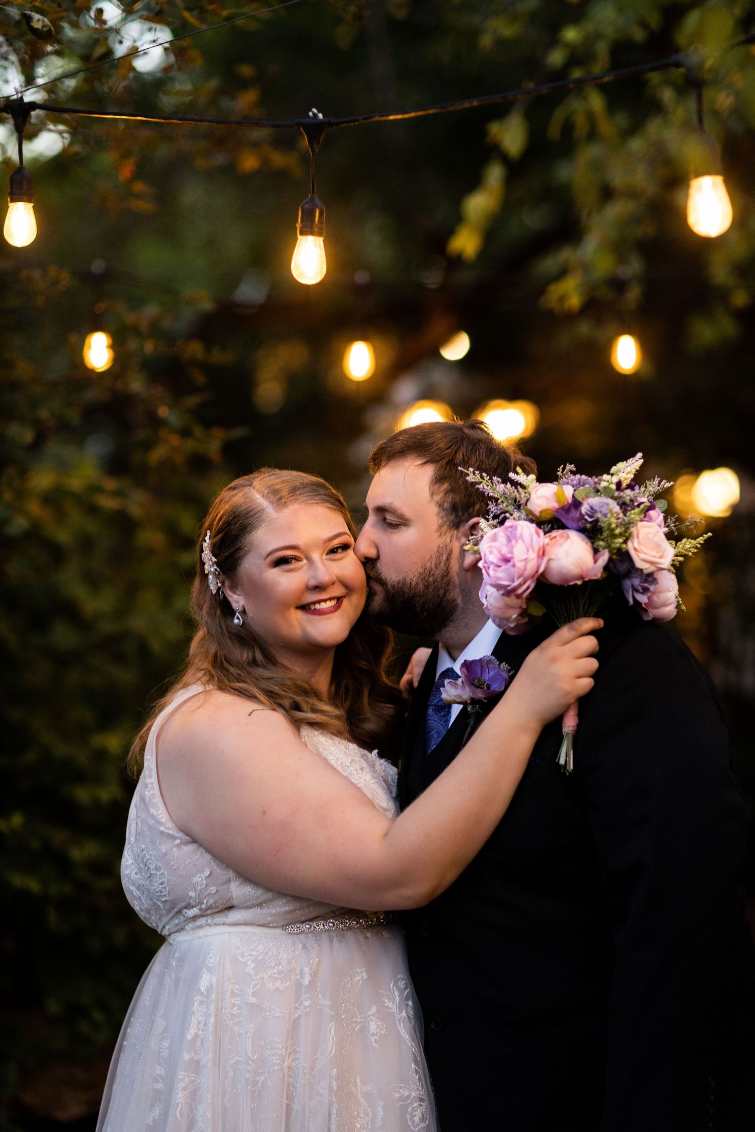 A romantic wedding moment captured under glowing string lights with a pink and purple floral bouquet.