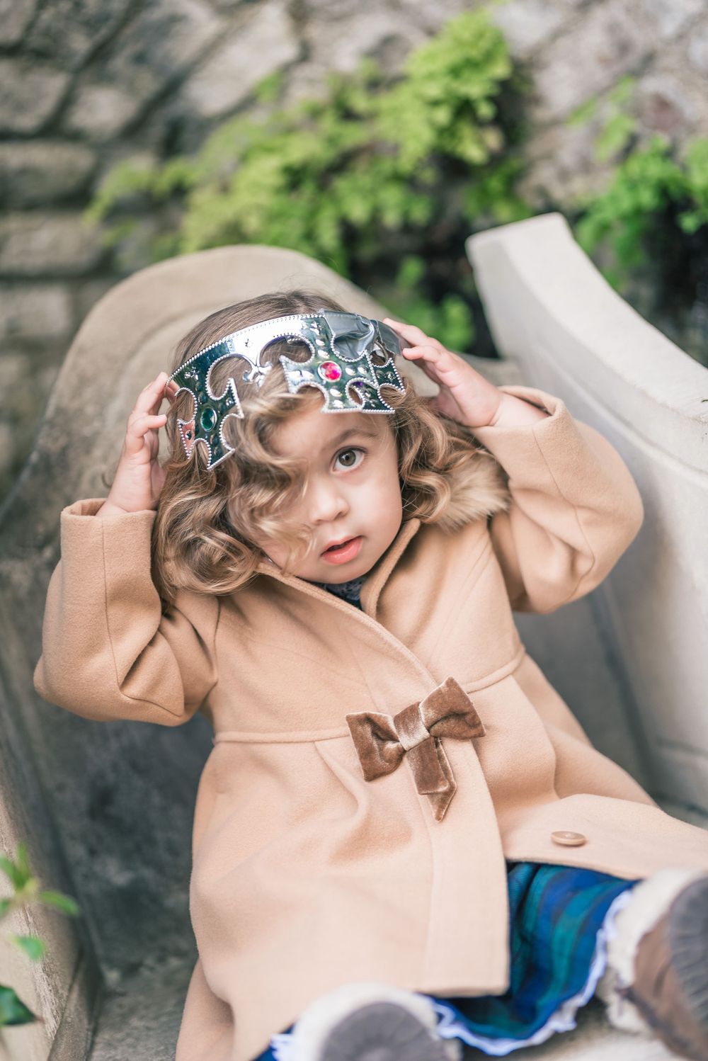 Curly-haired toddler in beige peacoat and decorative headpiece sits outdoors on stone steps.