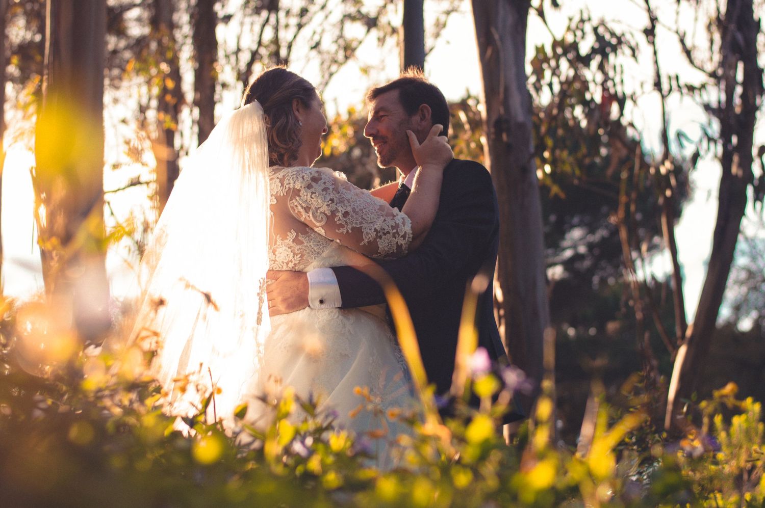 Romantic wedding couple embracing in sunlit forest wearing traditional white dress and formal attire.