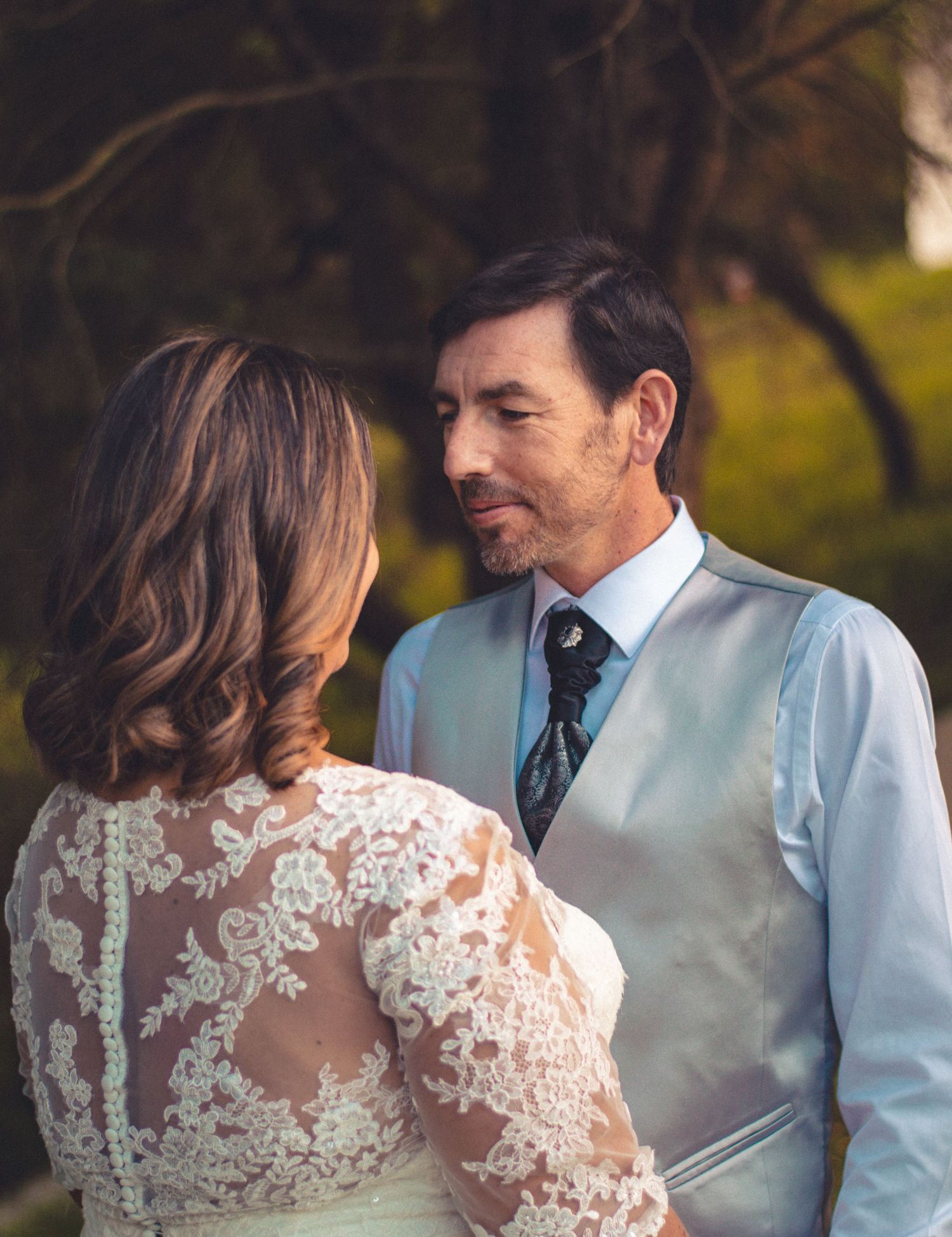Tender moment between bride in lace dress and groom in light vest during outdoor wedding.
